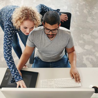 Top view shot of young man sitting at his desk working on pc and digital graphics tablet with woman pointing at monitor. Male graphic designer getting help from female coworker in office.